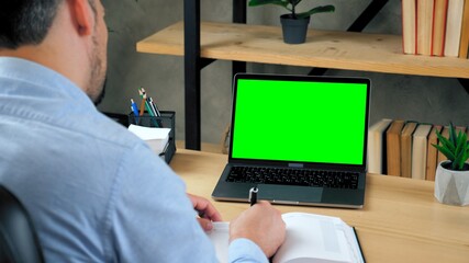 Over the shoulder. Businessman CEO manager at home office writes information with pen in notebook. Green screen laptop computer concept: online remote distance education by video call chat conference