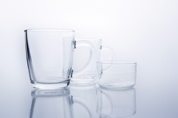 Two empty transparent glass mugs and saucer rosette on reflective surface on white background. Transparent glass still life.