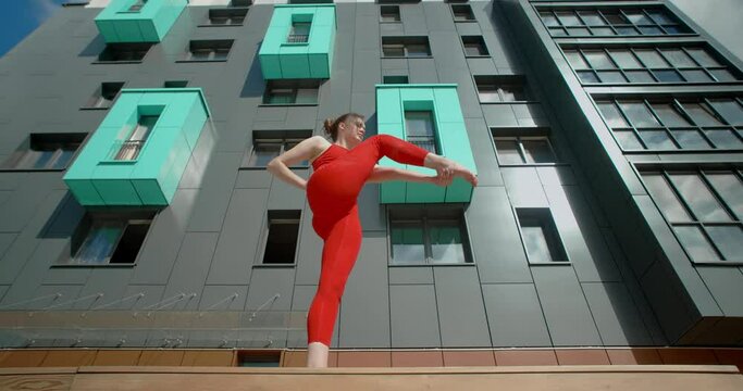 Woman in red does stretching and yoga exercices for legs on the bench in the yard of an apartment building, fitness in the urban environment, yoga class outdoors, physical workout in the city, 4k 120p