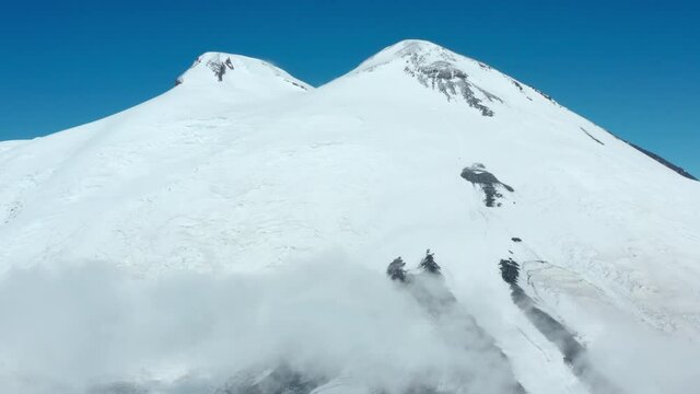 Snowy highest peak in the Caucasus Mountains of picturesque old high Elbrus with flowing fog clouds against clear blue sky on sunny day panoramic aerial view.