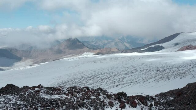 Red shelter station for skiers and tourists on snowy glacier ice cracks slope of Elbrus mountain under sky with clouds on sunny day aerial view.