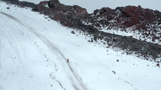 Climber in red jacket walks trekking up along track on steep snowy slope of old high Elbrus mountain on sunny cold day bird eye aerial view.