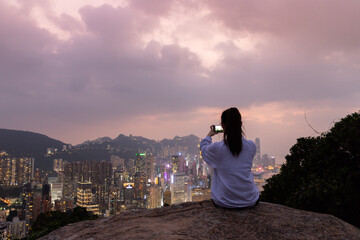 Silhouette of a girl who sits at rock and takes photos of sunset night view of city Skyline of Hong Kong Island (south of Victoria Harbour) from Braemar hill, North Point