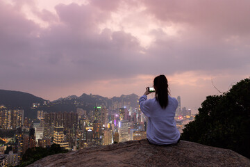 Fototapeta premium Silhouette of a girl who sits at rock and takes photos of sunset night view of city Skyline of Hong Kong Island (south of Victoria Harbour) from Braemar hill, North Point