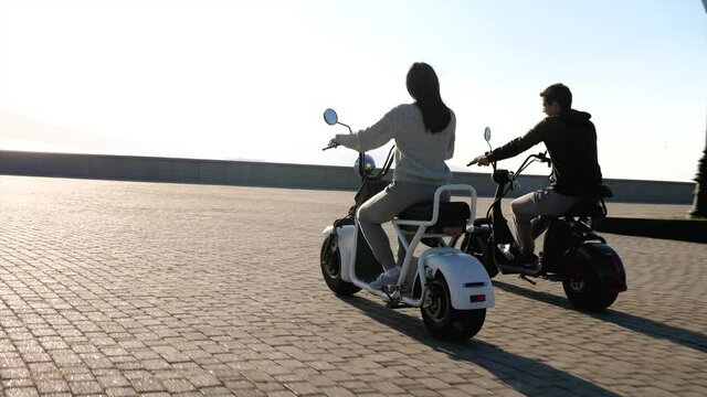 Man and girl on electric scooters are riding through urban seafront at morning. Happy couple is spending time while riding e-bikes next to each other. Ecological electro transport concept.