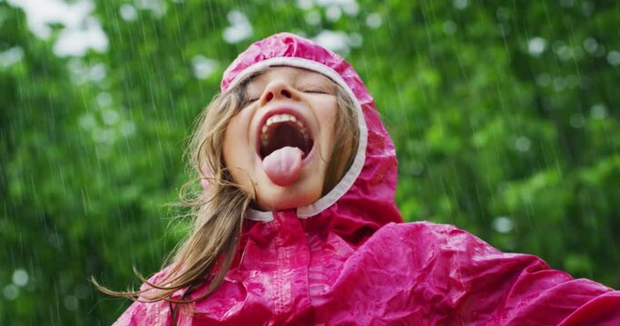 Authentic shot of carefree happy little girl in raincoat is having fun and enjoying the rain in a hot summer day in a green park. 