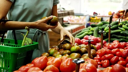 Unrecognizable caucasian woman with shopping basket is choosing green tomatoes in grocery store.