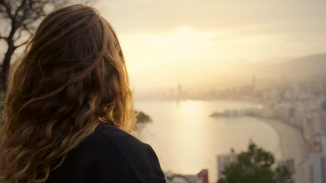 Young woman overlooks beautiful epic sunset over high rise buildings in city landscape. Futuristic modern city, woman with long hair contemplating and peaceful, look at horizon. Space for copy text