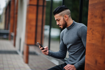 Portrait of stylish arab beard man wear grey turtleneck. Arabian model guy sit and look at smartphone.