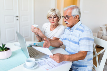 Happy aged husband and wife hold papers using laptop for online banking. Drinking coffee
