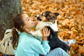 Woman holding her cute pupy Welsh Corgi dog, spending time together at autumn park with fallen leaves on background. Cute moments pets and human.