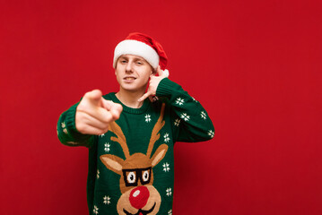 Funny young man in christmas clothes stands on a red background, looks into the camera, shows a call gesture and thumbs into the camera. Cheerful guy in santa hat shows call me gesture