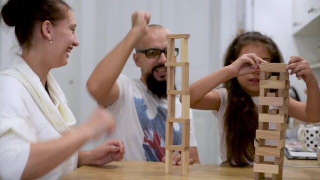 Happy dad and mom helping cute preschool little girl playing toys together on the table, building constructor tower of colorful wooden blocks at home.