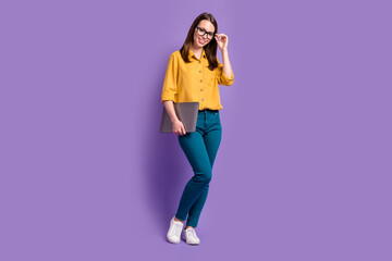 Full length photo of lady hold pc specs finger wear glasses yellow shirt blue trousers sneakers isolated purple color background