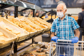 older european man wearing mask and gloves with covid protection chooses buns and bread in supermarket bakery
