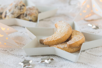 Fototapeta na wymiar Vanilla-flavored crescent cookies on a white star plate on white blanket, decorated with silver stars and starlight, mini stollen in the background