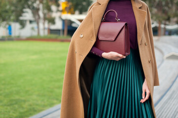 Fashionable young woman wearing green pleated midi skirt, sweater, high heel shoes, beige wool coat and holding burgundy handbag in hand on the city street. Street style.