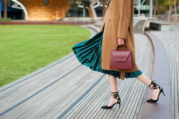 Fashionable young woman wearing green pleated midi skirt, sweater, high heel shoes, beige wool coat and holding burgundy handbag in hand on the city street. Street style.