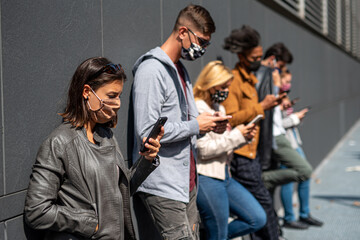 Fototapeta premium young Caucasian woman who reads and inquires about the corona virus infection in the foreground, group of multi-ethnic friends addicted to the use of cell phone, all wear protective masks, new normal