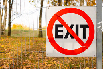 Prohibiting sign on a metal fence mesh. Red crossed out circle on a white background with the inscription EXIT. In the background there is a park with yellow foliage. Autumn day. Copy space.