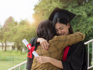 Asian female graduate wearing mortar board, holding certificate hugging senior woman tight with happiness.