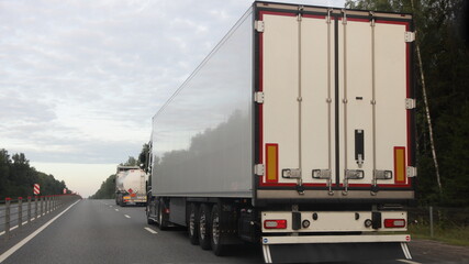 European road semi trucks with white trailer van and barrel move on two lane suburban asphalted highway motorway, rear view at summer evening on forest and sky background, delivery cargo logistics
