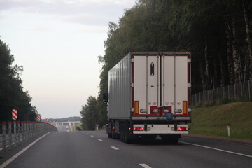 European long vehicle truck with white semi trailer van move on two lane suburban asphalted highway road, back view at summer evening on forest and sky background, carriage transportation logistics