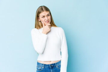 Young blonde woman isolated on blue background touching back of head, thinking and making a choice.