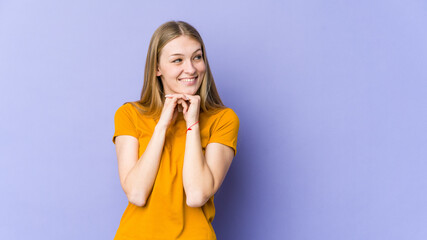 Young blonde woman isolated on purple background keeps hands under chin, is looking happily aside.