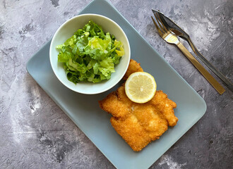 Breaded cod fish fillet, also known as Kabeljau. The fish is coated in egg, flour and breadcrumbs, then deep fried to golden brown and served with green salad with Vinaigrette and lemon.