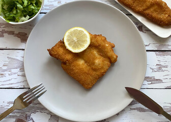 Breaded cod fish fillet, also known as Kabeljau. The fish is coated in egg, flour and breadcrumbs, then deep fried to golden brown and served with green salad with Vinaigrette and lemon.