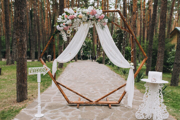 A wooden wedding arch decorated with flowers and white cloth stands in the forest among the pine...