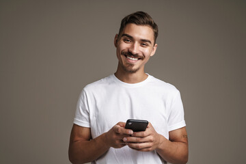 Image of joyful unshaven guy smiling and using cellphone