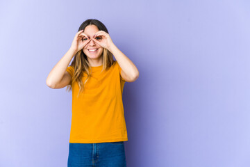 Young caucasian woman isolated on purple background showing okay sign over eyes