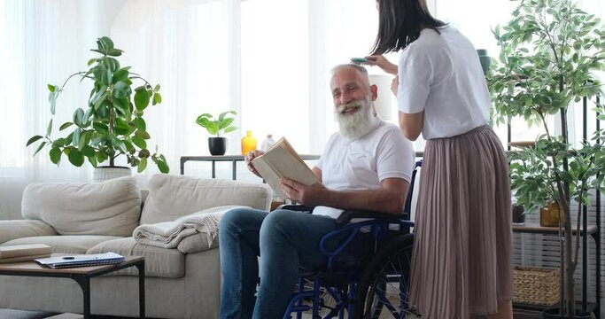 Young adult daughter combing hair of disabled old father reading book on wheelchair at home