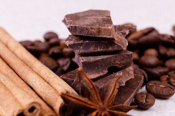 Stack of chocolate bar pieces with heap of roasted coffee beans and spices, cinnamon sticks and anise stars on a white table. Macro shot.