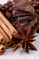 Stack of chocolate bar pieces with heap of roasted coffee beans and spices, cinnamon sticks and anise stars on a white table. Macro shot.