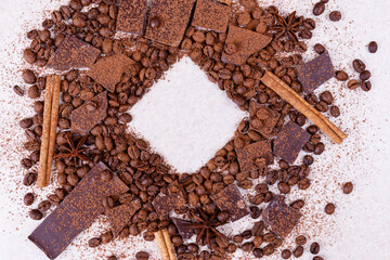 Pieces of chocolate bar on a white table with roasted coffee beans and spices, cinnamon sticks and anise stars, covered with cocoa powder. Copy space in the center in square form. Top view.