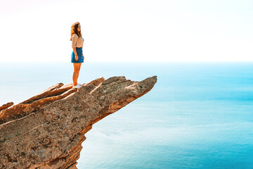 A young woman stands on a picturesque rock ledge above the sea against the sky. The concept of travel and freedom. 