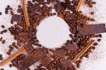 Pieces of chocolate bar on a white table with roasted coffee beans and spices, cinnamon sticks and anise stars, covered with cocoa powder. Copy space in the corner in circle form. Top view.