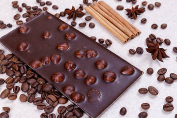 Chocolate bar with nuts on a white table with coffee beans and spices. Roasted coffee beans, cinnamon sticks and anise stars. Chocolate bar with hazelnuts. 