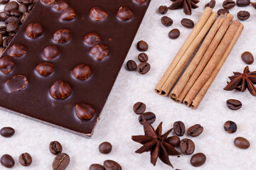 Chocolate bar with nuts on a white table with coffee beans and spices. Roasted coffee beans, cinnamon sticks and anise stars. Chocolate bar with hazelnuts. 
