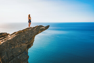 A young woman stands on a picturesque rock ledge above the sea against the sky. The concept of travel and freedom.  - Powered by Adobe