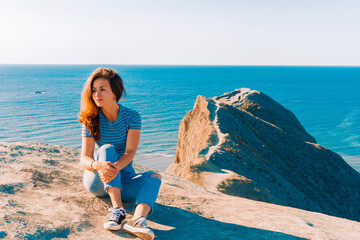 A young woman with lonh hair  sits on the slope of Cape chameleon with an amazing panoramic view of the hills and the sea from above