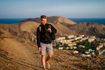 A young blond man with a backpack walks along a mountain path with a view of the coastal city between the hills, Crimea