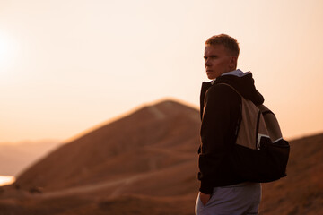 Picturesque mountain landscape at sunset with the sea on the horizon, rear view of young blond man with backpack stands on the slope in the orange rays of the sunset