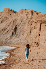 A young brunette woman in jeans runs along a picturesque sandy beach with a view of the Cape Chameleon rock in Crimea