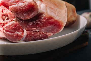 Obraz premium Raw pork knuckle on white plate on grey wooden background, close-up. Cooking of meat dish.