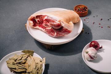 Raw pork knuckle on white plate, seasonings, garlic, red onions, pepper set, bay leaf on grey wooden background. Cooking of meat dish, meal and flavourings, cuisine.