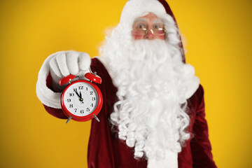 Santa Claus holding alarm clock on yellow background, focus on hand. Christmas countdown
